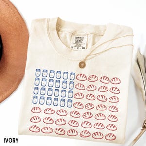 May include: Ivory-colored t-shirt with a graphic design resembling the American flag. The design features blue jars in the place of stars and red bread loaves in place of stripes. A gold necklace is draped across the shirt.