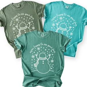 May include: Three short-sleeved t-shirts in shades of green and blue. Each shirt features a white graphic of a snowman with a scarf and snowflakes, and the text "snow happy to see your face".