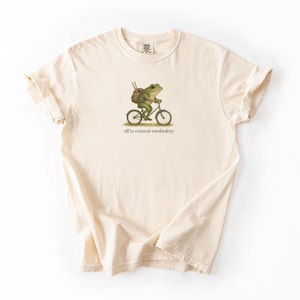 May include: Cream-colored t-shirt featuring a green frog riding a bicycle with a backpack. The frog illustration is above the text "off to commit tomfoolery". The shirt has rolled-up sleeves and a tag at the neckline.