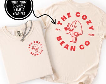 May include: Two cream-colored t-shirts with red graphics. One shirt has a front logo of a bean character and the text "THE COZY BEAN CO." The other shirt has the same logo on the back and the text "THE COZY BEAN CO. EST. 2025". A silver chain necklace is on the folded shirt.