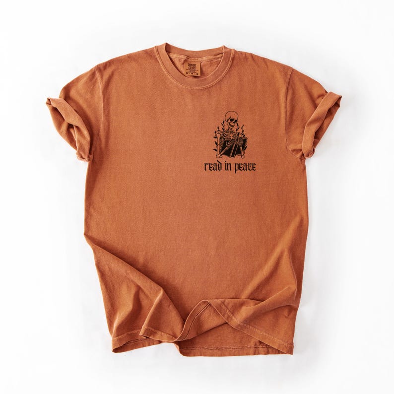 Puede incluir: Camiseta de color naranja quemado con un gr&aacute;fico negro de un esqueleto leyendo un libro, rodeado de llamas. El texto "read in peace" est&aacute; debajo. La camiseta tiene cuello redondo y mangas cortas.