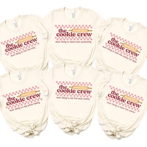 May include: Set of five cream-colored t-shirts with pink and white checkered patterns and the text "the cookie crew" in red and brown. Each shirt features a rolling pin and a different phrase about cookies.