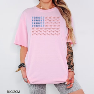 May include: A light pink t-shirt featuring a graphic design of an American flag made of hot dogs and blue drinks. The shirt has short sleeves and a relaxed fit. The word "BLOSSOM" is printed at the bottom left.