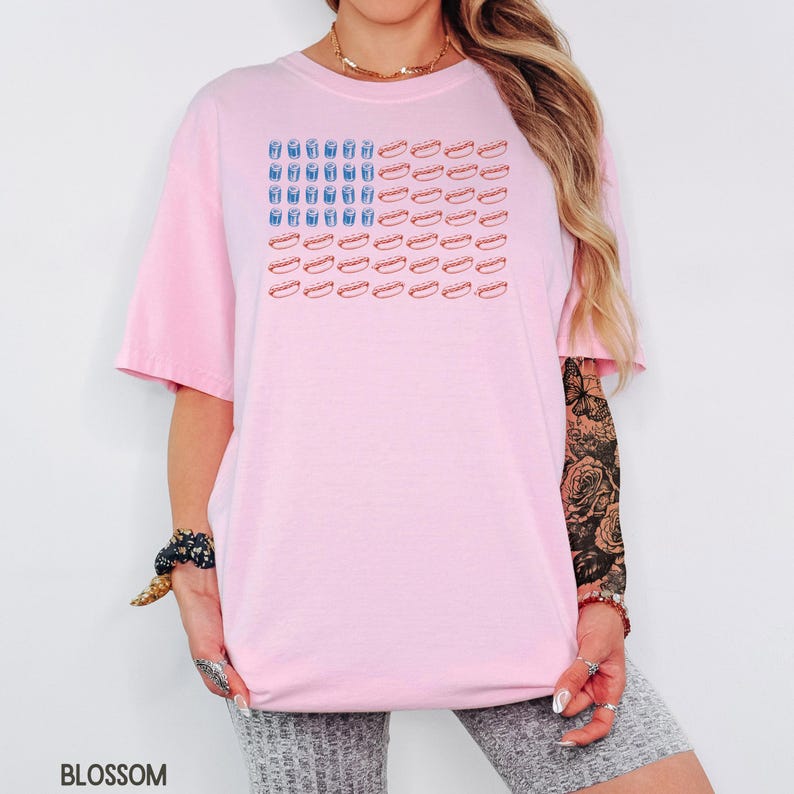 May include: A light pink t-shirt featuring a graphic design of an American flag made of hot dogs and blue drinks. The shirt has short sleeves and a relaxed fit. The word "BLOSSOM" is printed at the bottom left.