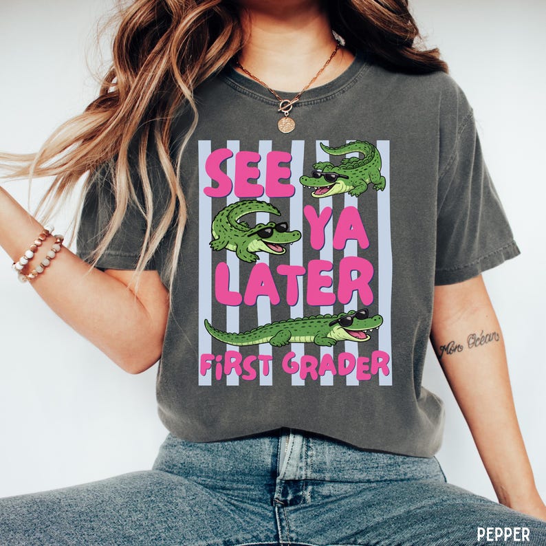 K&ouml;nnte beinhalten: Ein anthrazitfarbenes Kurzarm-T-Shirt mit einem Grafikdruck von drei gr&uuml;nen Alligatoren mit schwarzer Sonnenbrille. Der Text "SEE YA LATER" und "FIRST GRADER" ist in rosa Buchstaben auf hellblauen vertikalen Streifen abgebildet.