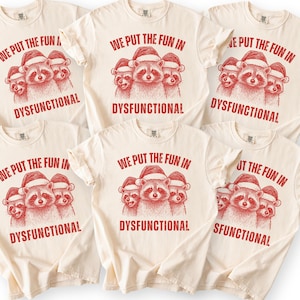 May include: A cream-colored short-sleeve t-shirt featuring a red graphic of three raccoons wearing Santa hats. The text above the raccoons reads WE PUT THE FUN IN and the text below reads DYSFUNCTIONAL. The design is printed in a distressed red ink.