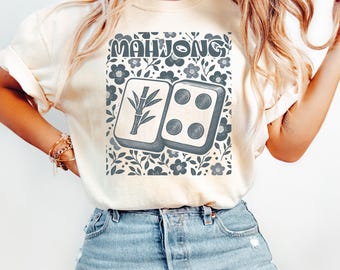 May include: Cream-colored t-shirt featuring a graphic design with the word "MAHJONG" above two Mahjong tiles. The tiles are gray with bamboo and dot symbols, set against a floral background. A casual, stylish top.
