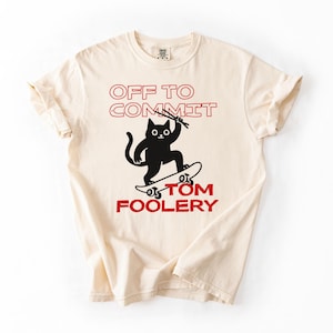 May include: Cream-colored t-shirt with the text "OFF TO COMMIT TOM FOOLERY" in red. A black cat is skateboarding, holding two sticks. The shirt has short sleeves and a crew neck.