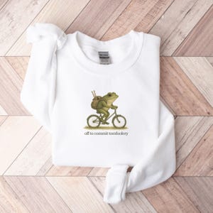 May include: White sweatshirt featuring a green frog riding a bicycle with a backpack. The frog illustration is accompanied by the text "off to commit tomfoolery". The sweatshirt is made of a soft material.