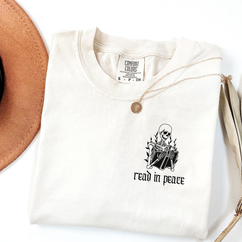 Puede incluir: Camiseta color crema con un gr&aacute;fico de esqueleto negro leyendo un libro, acompa&ntilde;ada de las palabras "read in peace". Tambi&eacute;n se ven un collar dorado y un sombrero marr&oacute;n. La camiseta est&aacute; doblada cuidadosamente.