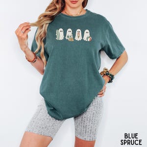 May include: A blue spruce colored short-sleeve t-shirt featuring a graphic print of five white ghosts engaged in beach activities, including holding a surfboard, a bucket, a sandcastle, an ice cream cone, and a beach ball, centered on the chest.