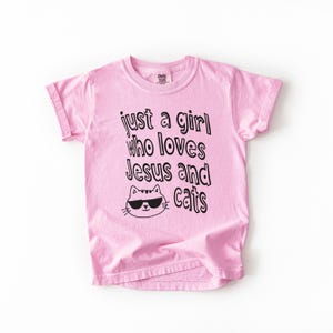May include: Pink t-shirt with the text "just a girl who loves Jesus and cats" in black. A cartoon cat wearing sunglasses is below the text. The shirt has short sleeves and a crew neck.