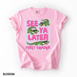 K&ouml;nnte beinhalten: Ein hellrosa T-Shirt mit kurzen &Auml;rmeln und einer Grafik von drei gr&uuml;nen Alligatoren mit schwarzer Sonnenbrille. Der Text lautet "SEE YA LATER" in gro&szlig;en rosa Buchstaben und "FIRST GRADER" in kleineren rosa Buchstaben auf hellblauen vertikalen Streifen.