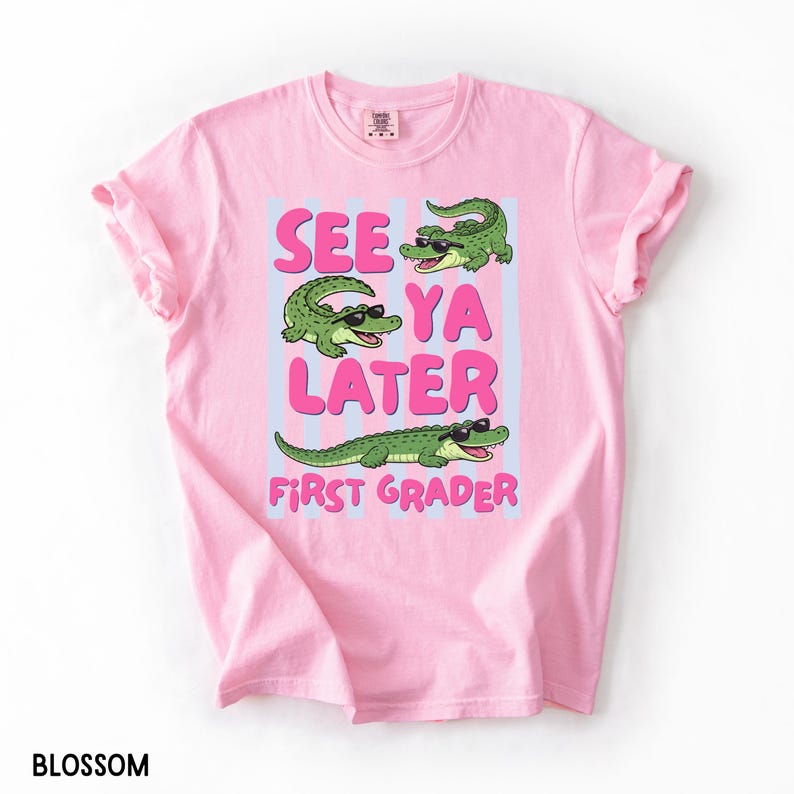 K&ouml;nnte beinhalten: Ein hellrosa T-Shirt mit kurzen &Auml;rmeln und einer Grafik von drei gr&uuml;nen Alligatoren mit schwarzer Sonnenbrille. Der Text lautet "SEE YA LATER" in gro&szlig;en rosa Buchstaben und "FIRST GRADER" in kleineren rosa Buchstaben auf hellblauen vertikalen Streifen.