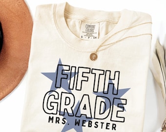 Peut inclure: T-shirt ivoire avec l'inscription "FIFTH GRADE MRS. WEBSTER" en noir sur un motif d'éclat d'étoile bleu. Un collier doré avec un pendentif repose sur le t-shirt. Un chapeau marron est partiellement visible.