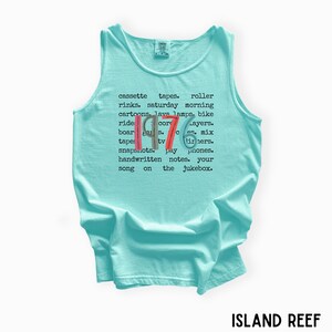 K&ouml;nnte beinhalten: Ein hellblaues Tanktop mit dem Wort "1976" in Rot, Wei&szlig; und Blau. Das Shirt zeigt eine Liste nostalgischer Gegenst&auml;nde und Phrasen aus den 1970er Jahren, wie Kassetten, Lavalampe und Rollschuhbahnen. Der Text "ISLAND REEF" befindet sich unten.