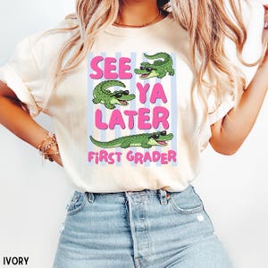 K&ouml;nnte beinhalten: Ein elfenbeinfarbenes T-Shirt mit einem Grafikdruck von drei gr&uuml;nen Alligatoren mit schwarzer Sonnenbrille. Der Text lautet "SEE YA LATER" in gro&szlig;en rosa Buchstaben und "FIRST GRADER" in kleineren rosa Buchstaben vor hellblauen Streifen.
