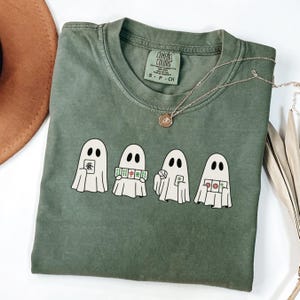 May include: Green Comfort Colors t-shirt featuring four cartoon ghosts holding Mahjong tiles. The shirt has a crew neck and short sleeves. A silver necklace with a small pendant is draped across the shirt.