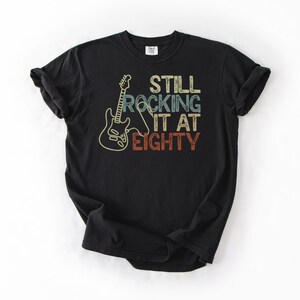May include: Black t-shirt with a guitar graphic and the text "Still Rocking It At Eighty" in beige, blue, and red. The shirt is made of a soft material and is suitable for casual wear.
