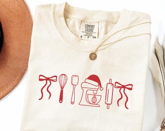 May include: Cream-colored Comfort Colors t-shirt with a red graphic of baking tools and a Santa hat. A gold necklace with a pendant is draped across the shirt. The shirt is folded on a white surface.