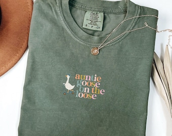 May include: An olive green short-sleeve t-shirt with a crew neckline. The chest features an embroidered graphic of a white goose on a skateboard with the multi-colored text "auntie goose on the loose". The inner neck label reads Comfort Colors.