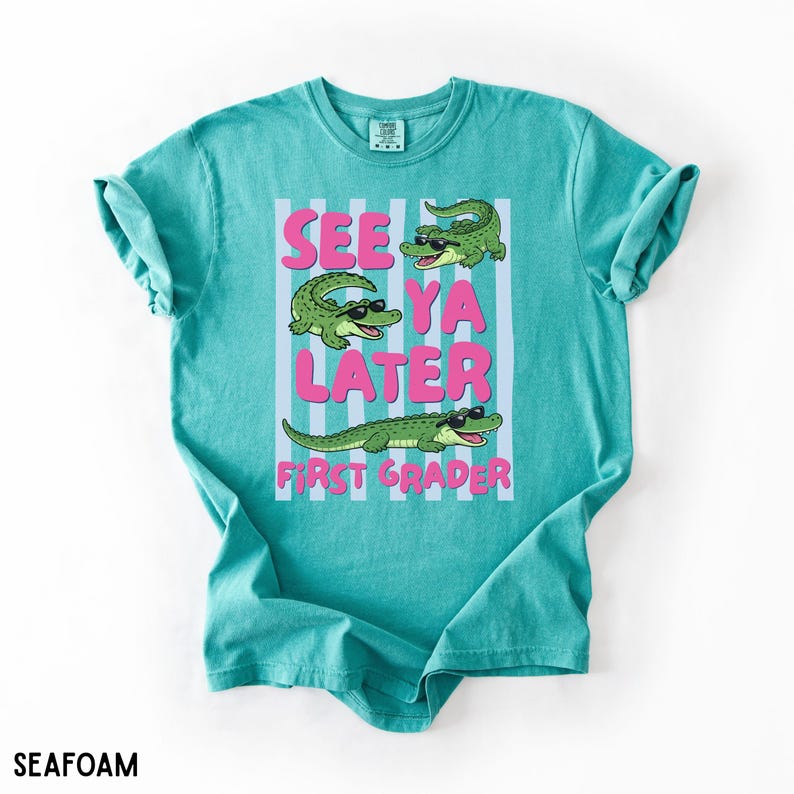 K&ouml;nnte beinhalten: Ein meergr&uuml;nes T-Shirt mit einem Grafikdruck von drei gr&uuml;nen Alligatoren, die eine schwarze Sonnenbrille tragen. Der Text "SEE YA LATER FIRST GRADER" steht in fetten rosa Buchstaben auf einem Hintergrund aus hellblauen und wei&szlig;en Streifen.