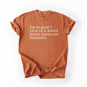 May include: A rust-colored t-shirt with the text "I'm so glad I live in a world where there are Octobers" printed in white. The shirt is folded and displayed on a white background.