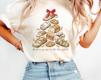 May include: Cream-colored t-shirt featuring a Christmas tree design made of various types of bread, adorned with a red bow and string lights. The phrase "baking spirits bright" is written in a script font below the tree.