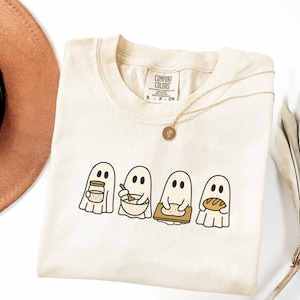 May include: A cream-colored t-shirt featuring a graphic of four white ghosts engaged in baking activities. The ghosts are shown holding a jar, mixing in a bowl, kneading dough on a board, and holding a loaf of bread. The shirt has a Comfort Colors label.
