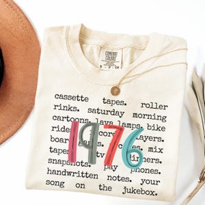 May include: Cream-colored t-shirt with the year "1976" in colorful block letters. The shirt features a list of nostalgic words like "cassette tapes" and "roller rinks". A gold necklace with a pendant rests on the shirt.