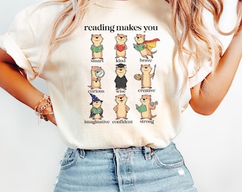 May include: A cream-colored t-shirt featuring a graphic of nine otters with the text reading makes you. Each otter is labeled with a trait: smart, kind, brave, curious, wise, creative, imaginative, confident, and strong, depicted with related items.
