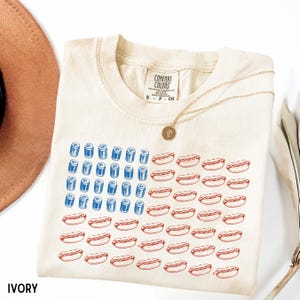 May include: Ivory-colored t-shirt with a graphic design of the American flag made of blue cans and red hot dogs. The shirt has a crew neck and short sleeves. The brand label "Comfort Colors" is visible. A gold necklace with a pendant is draped on the shirt.