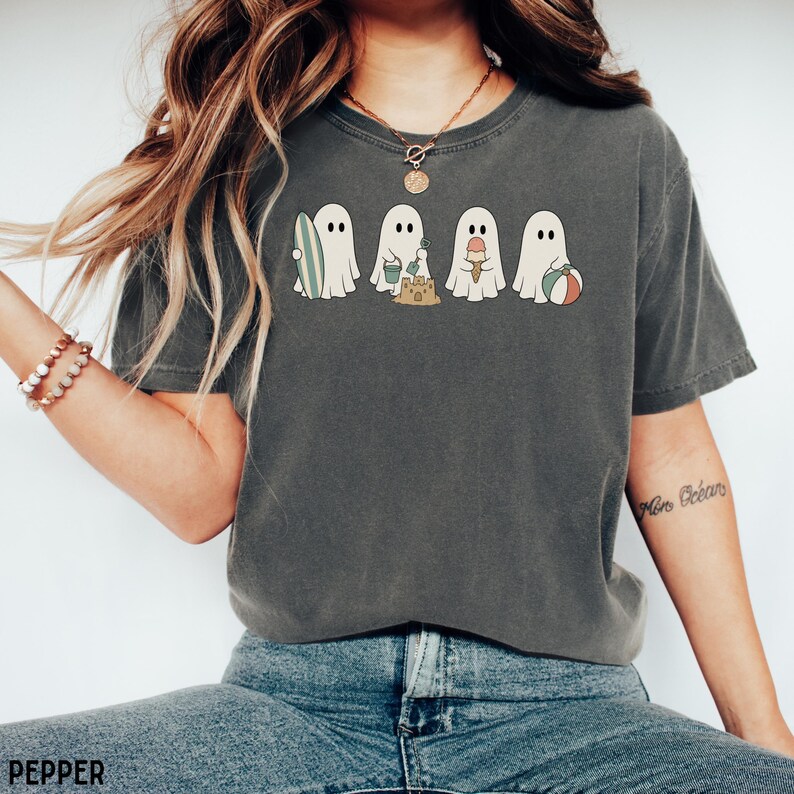 May include: A charcoal gray short-sleeve t-shirt featuring a graphic print of four white ghosts engaged in beach activities. One ghost holds a surfboard, another builds a sandcastle, the third holds an ice cream cone, and the fourth holds a beach ball.