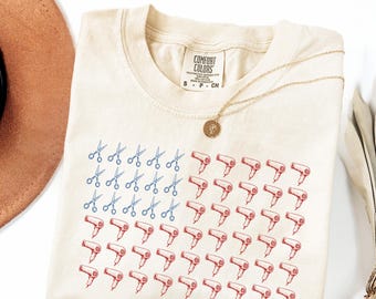 May include: An ivory t-shirt featuring a graphic print arranged like an American flag, with blue scissors in the upper left corner and red hair dryers forming the stripes. The shirt has a Comfort Colors label at the neck and a gold pendant necklace.