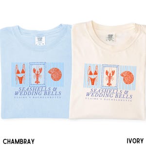 May include: Two crew neck t-shirts in chambray blue and ivory, each featuring a graphic print of a bikini, a lobster, and a seashell over blue vertical stripes. The text reads SEASHELLS & WEDDING BELLS and CLAIRE'S BACHELORETTE in blue lettering.