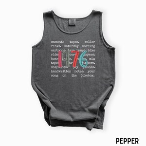 K&ouml;nnte beinhalten: Ein dunkelgraues Tanktop mit dem Wort "1976" in Rot, Blau und Gr&uuml;n. Das Tanktop hat eine Liste von W&ouml;rtern in Wei&szlig;, darunter "Kassetten," "Rollschuhbahnen" und "Cartoons."