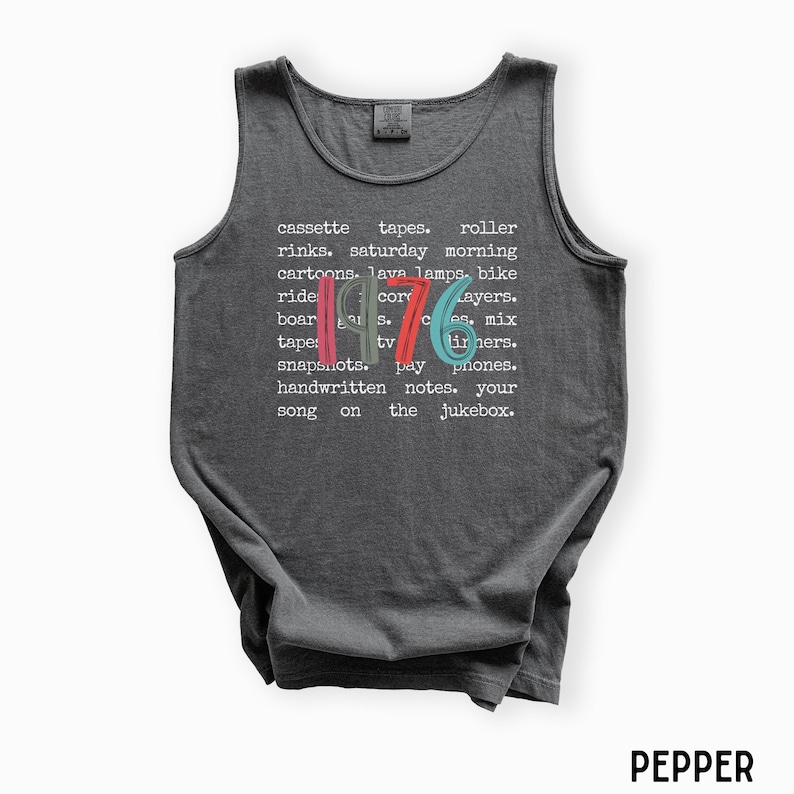 K&ouml;nnte beinhalten: Ein dunkelgraues Tanktop mit dem Wort "1976" in Rot, Blau und Gr&uuml;n. Das Tanktop hat eine Liste von W&ouml;rtern in Wei&szlig;, darunter "Kassetten," "Rollschuhbahnen" und "Cartoons."