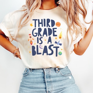 May include: A cream-colored t-shirt featuring the dark blue text "THIRD GRADE IS A BLAST" surrounded by colorful space-themed graphics including a rocket ship, a planet, a satellite, a comet, a crescent moon, and several yellow stars.