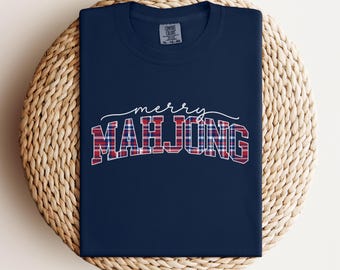 May include: Navy blue t-shirt with the words "merry MAH JONG" in white script and plaid lettering. The shirt is folded on a woven, circular surface. The shirt is a Comfort Colors brand.