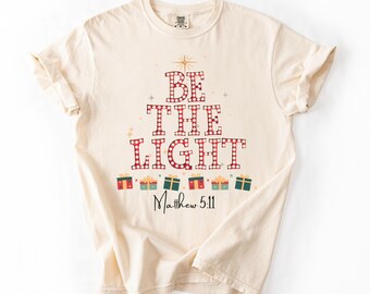 May include: Cream-colored t-shirt with the words "BE THE LIGHT" in a red and white checkered pattern. Below the text are colorful gift box graphics. The text "Matthew 5:11" is printed in black script.