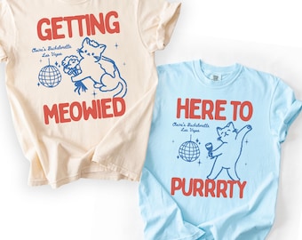 Op de afbeelding: Een ivoorkleurig t-shirt met rode tekst GETTING MEOWIED en een blauwe kat-afbeelding met discobal, en een chambray blauw t-shirt met rode tekst HERE TO PURRRTY en een blauwe kat-afbeelding met discobal, beide met de tekst Claire's Bachelorette Las Vegas.
