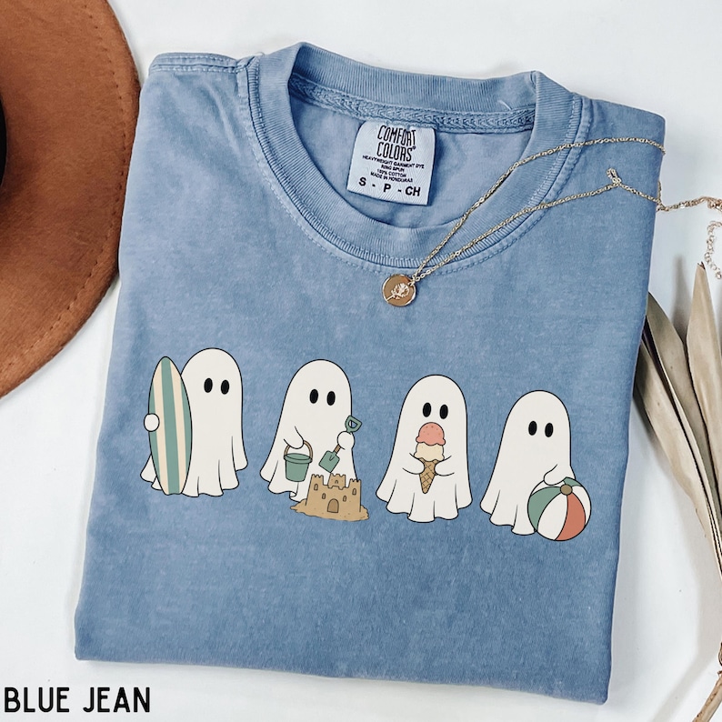 May include: A blue denim-colored t-shirt featuring a graphic of four white ghosts engaged in beach activities, including holding a surfboard, building a sandcastle, eating an ice cream cone, and holding a beach ball. The shirt has a Comfort Colors label.
