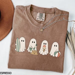 May include: An espresso-colored short-sleeve t-shirt featuring a graphic of four white ghosts at the beach. One ghost holds a surfboard, one builds a sandcastle with a bucket and shovel, one holds an ice cream cone, and one holds a beach ball.