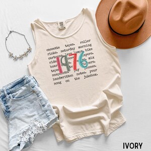 K&ouml;nnte beinhalten: Elfenbeinfarbenes Tanktop mit dem Jahr "1976" in bunten Buchstaben. Das Shirt zeigt eine Liste von W&ouml;rtern wie "Kassettenb&auml;nder", "Cartoons" und "Schnappsch&uuml;sse". Ein brauner Hut, Jeansshorts und eine Blumenkette sind ebenfalls im Bild.