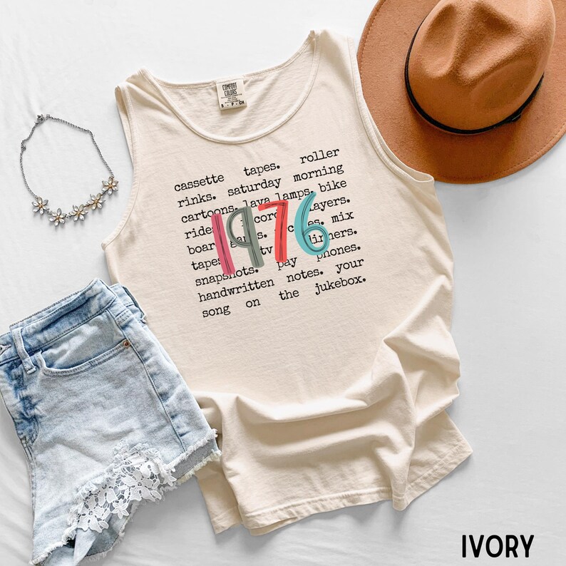 K&ouml;nnte beinhalten: Elfenbeinfarbenes Tanktop mit dem Jahr "1976" in bunten Buchstaben. Das Shirt zeigt eine Liste von W&ouml;rtern wie "Kassettenb&auml;nder", "Cartoons" und "Schnappsch&uuml;sse". Ein brauner Hut, Jeansshorts und eine Blumenkette sind ebenfalls im Bild.