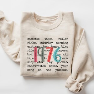 May include: Cream-colored sweatshirt with the year "1976" in colorful text. The sweatshirt features a list of nostalgic items and phrases from the 1970s, including cassette tapes, roller rinks, and lava lamps. A cozy and retro-themed apparel item.