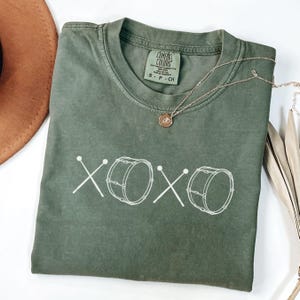 May include: An olive green t-shirt with the text "xoxo" and drum graphics in white. The shirt has a crew neck and short sleeves. A gold necklace with a pendant is draped across the shirt.