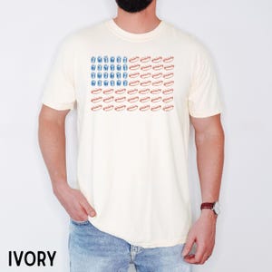 May include: Ivory-colored t-shirt featuring a graphic design resembling the American flag, with blue drink illustrations replacing the stars and red hot dogs replacing the stripes. The shirt is a casual, short-sleeved style.