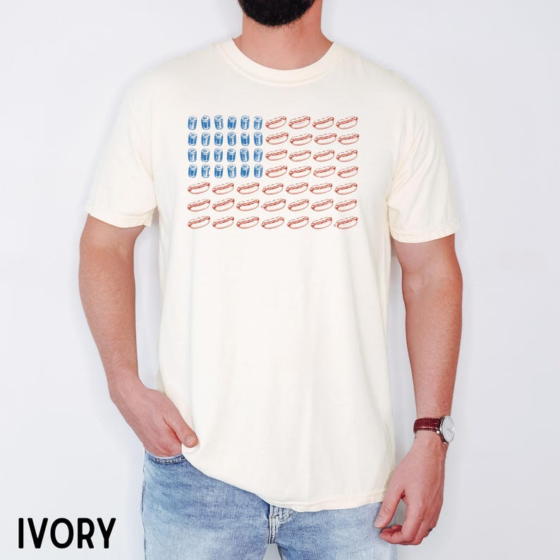 May include: Ivory-colored t-shirt featuring a graphic design resembling the American flag, with blue drink illustrations replacing the stars and red hot dogs replacing the stripes. The shirt is a casual, short-sleeved style.