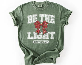 May include: Green t-shirt with the text "BE THE LIGHT" in white block letters. A red bow with Christmas lights is in the center. The text "MATTHEW 5:14" is below the bow.